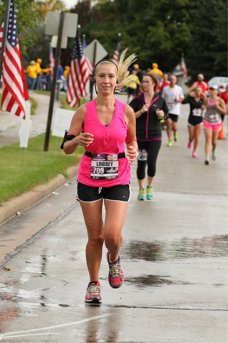 Lindsey is pictured wearing a hot pink running top, black running shorts, and blank and pink running shoes. The action shot shows her running a half marathon with other runners behind her.