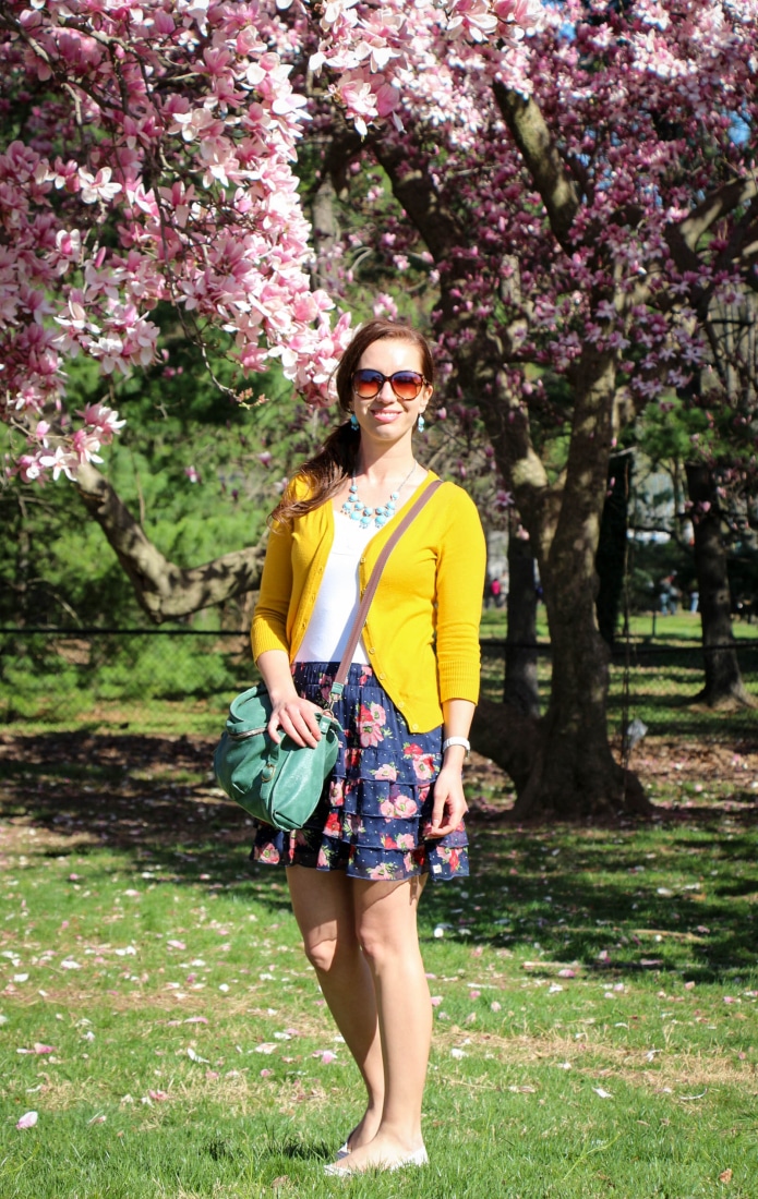 Lindsey poses in front of blooming cherry blossom trees. She's wearing a yellow cardigan over a white top paired with a navy and pink floral mini skirt.