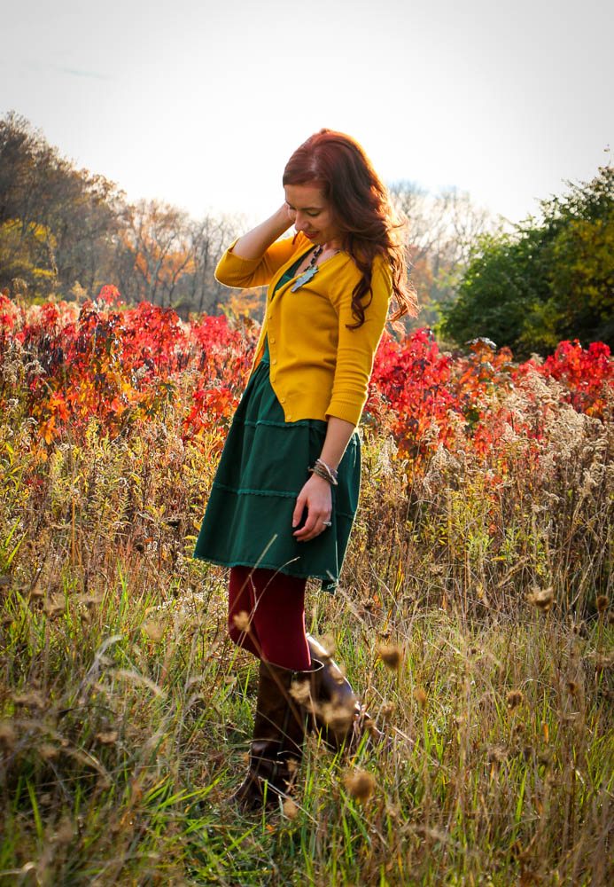 Lindsey poses in a colorful field filled with fall-colored foliage. She's wearing a yellow cardigan, emerald green skirt and dark red tights paired with knee-high boots.