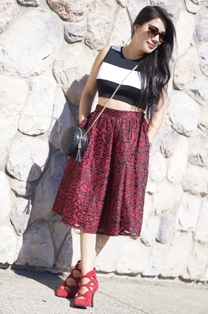 A woman poses against a stone wall. She's wearing a cropped black and white stripped shirt and red patterned knee-length skirt. The outfit is accessorized with sunglasses, a cross-body bag, and strappy red heels.