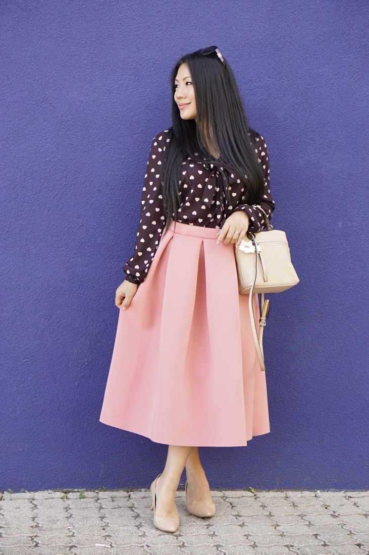 A woman stands in front of a blue-painted wall. She's wearing a black polka-dot blouse and light pink knee-length skirt paired with matching nude heels and a handbag.