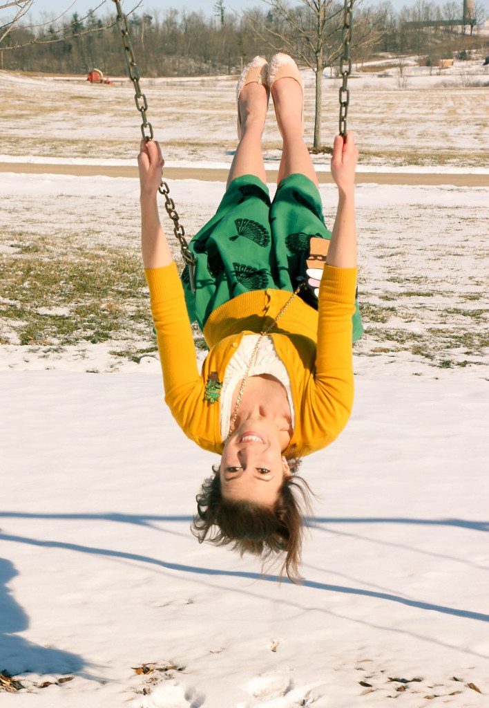 An image of Lindsey swinging on a swing set, hanging almost upside down. She's wearing a yellow cardigan paired with a green skirt and nude heels.