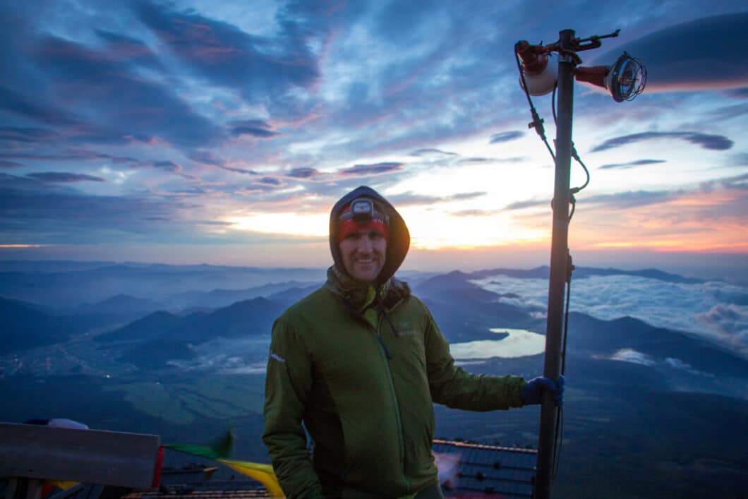 A man stands at a vantage point while hiking Mount Fiji wearing a heavy duty hiking jacket, headlamp, and hat.