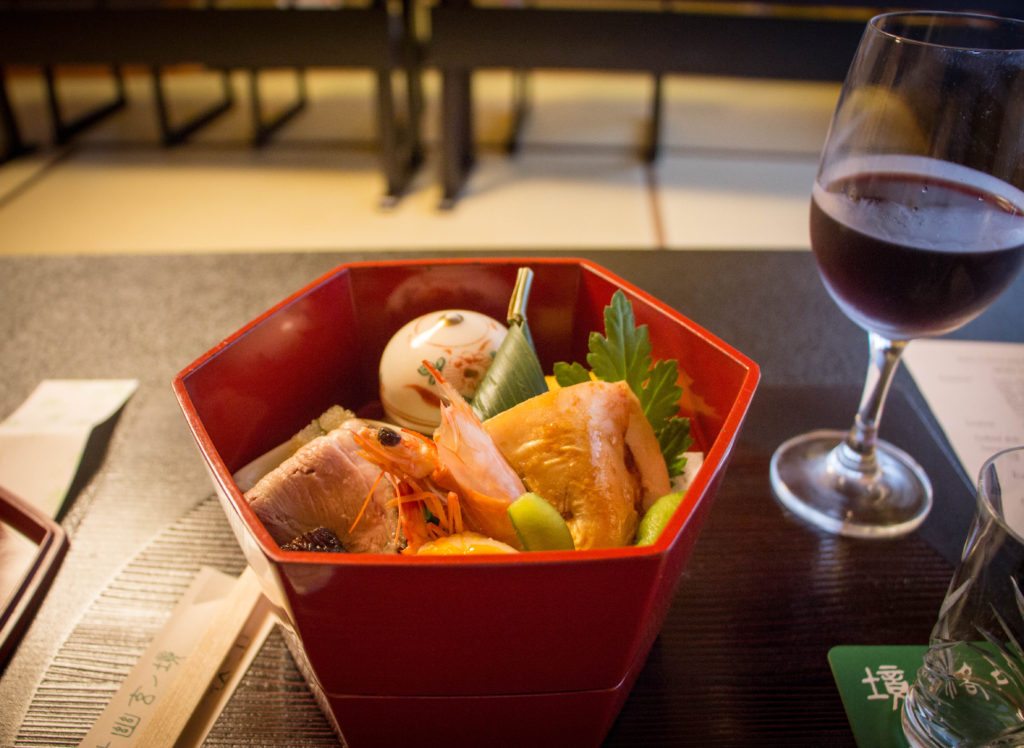 A red hexagon-shaped bowl holds pieces of seafood as part of a traditional Kaiseki dinner. There is also a glass of red wine on the table.