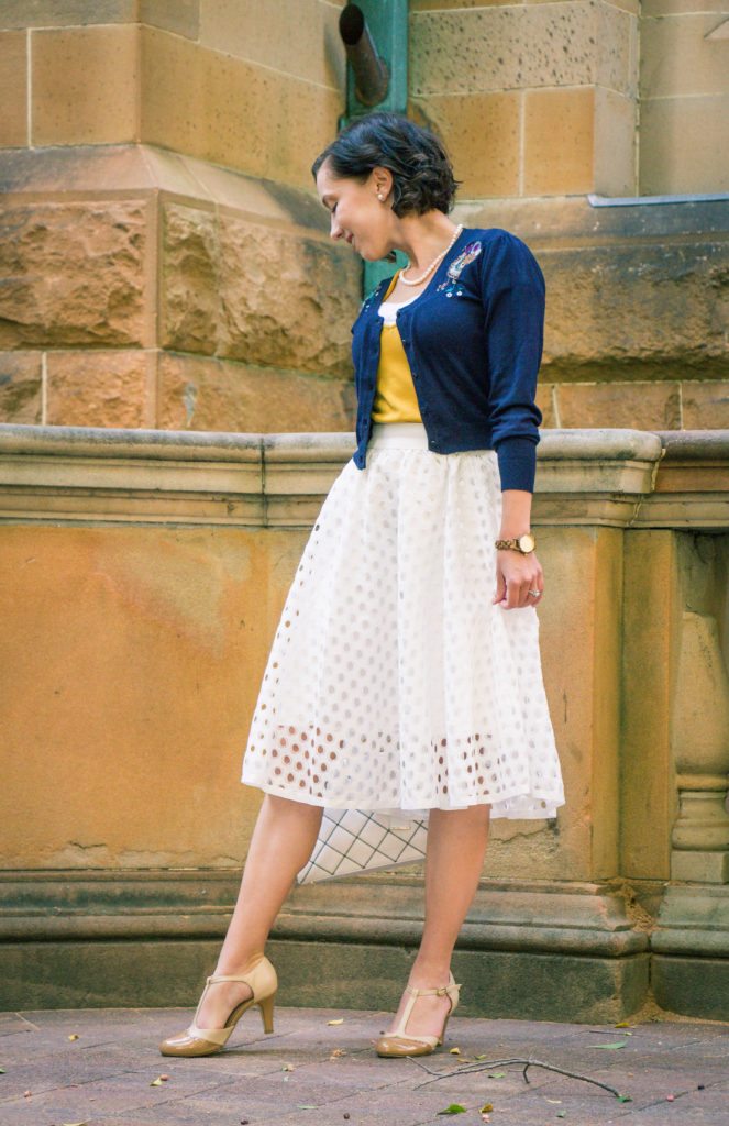 A woman poses in front of a large stone building while wearing a vintage inspired outfit. She has on a navy blue cardigan over a golden-yellow top and a white knee-length skirt and tan heels.