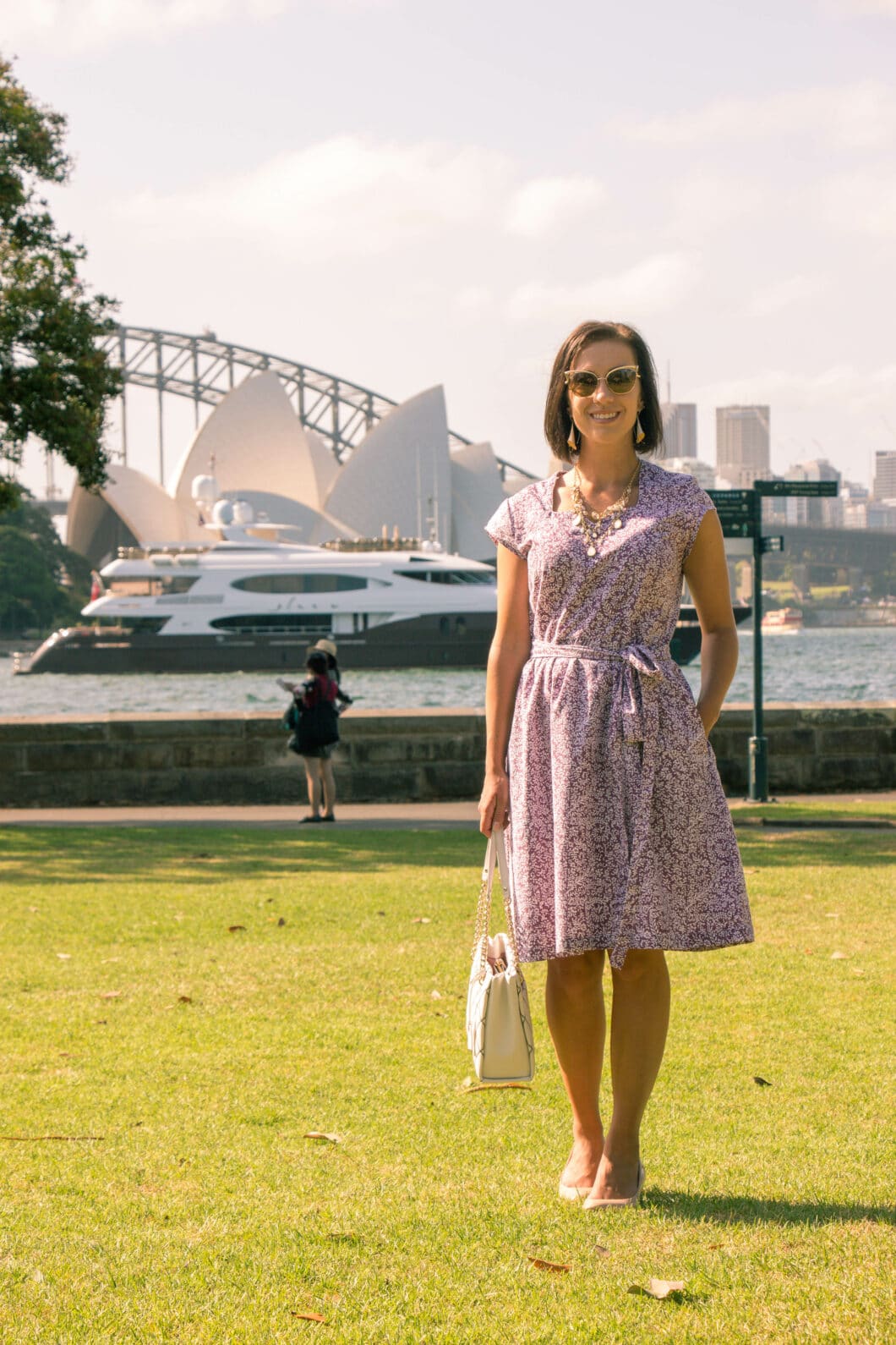 A woman stands in a park, the Sydney Opera House visible across the water. She's wearing a summery floral dress from sustainable travel clothing brand Passion Lillie.