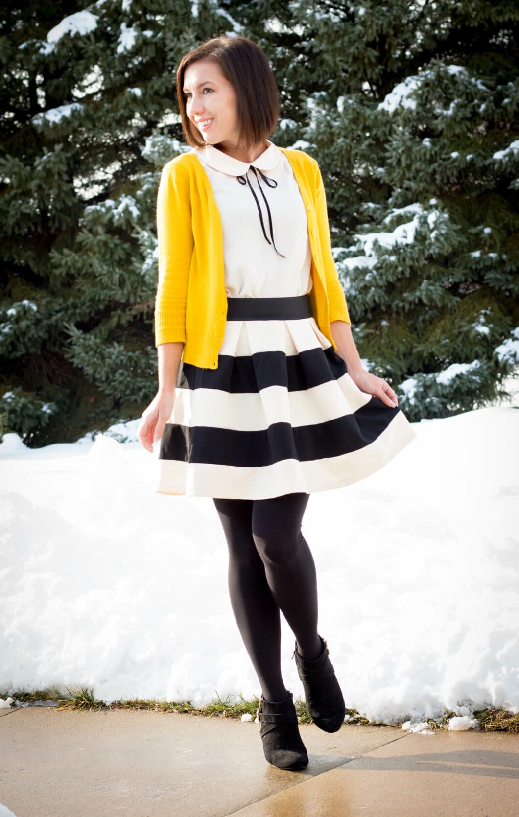 Lindsey poses in front of snowy trees. She's wearing a black and white striped shirt paired with a white top, yellow cardigan, and black tights and ankle boots.