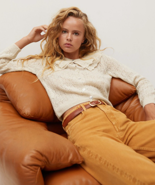 A young woman sits on a leather sofa wearing a Openwork Baby doll neck cardigan with mustard yellow corduroy pants from Mango 