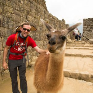 Zac wearing a red shirt and grey Eddie Bauer travel pants posing with a llama at Machu Picchu