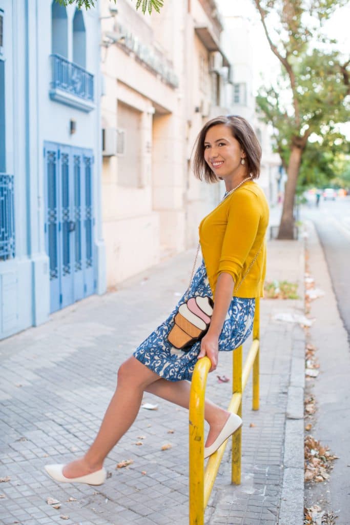 Lindsey poses sitting on a yellow guardrail. She's wearing a blue and white skater dress, yellow cardigan, and an over the shoulder purse shaped like an ice cream cone.