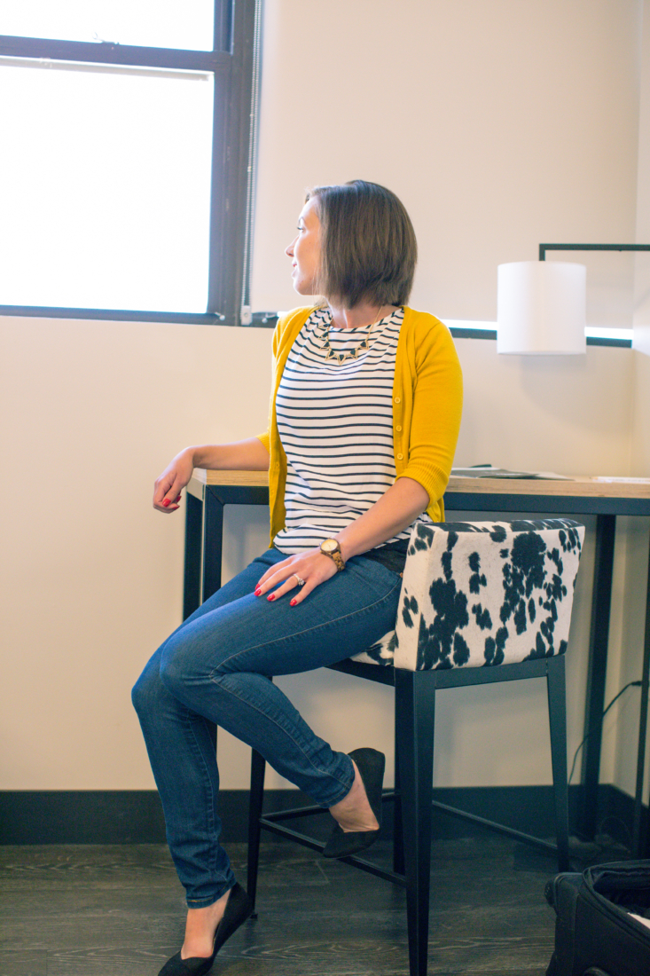 Lindsey poses sitting on a chair at a small desk. She's wearing jeans, a black and white striped shirt, and a yellow cardigan. She's looking over her shoulder out the window.