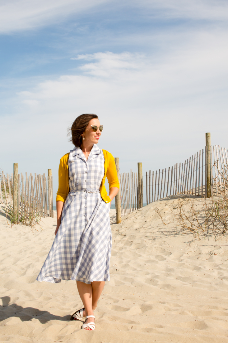 Lindsey poses on a sandy peach on a sunny, breezy day. She's wearing a vintage style light blue and white gingham dress paired with a yellow cardigan.