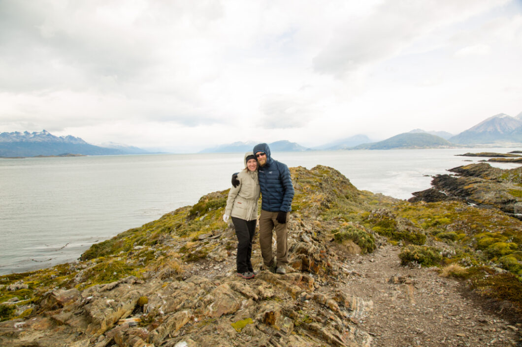 Zac and Lindsey of Have Clothes, Will Travel bundled up in warm clothing standing on Bridges Island, Ushuaia with the sea in the background