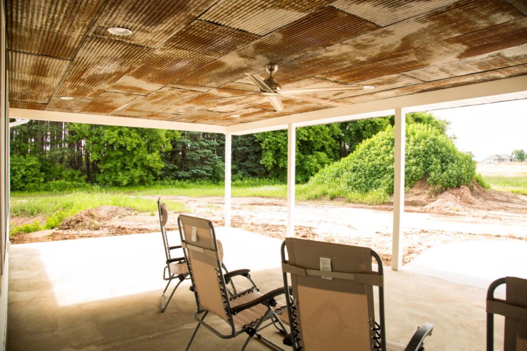 An empty backyard patio space filled with nothing but a few lawn chairs. The landscaping in the backyard is scarce, with a large grassy dirt hill in the center of the lawn.