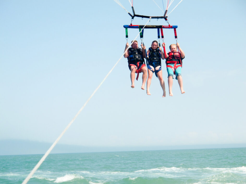 Three people are strapped into a parasailing harness, being towed by a rope. The people hover above the turquoise-colored water and sky. 