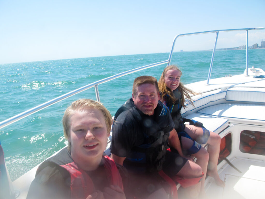 Three people are sitting on the side of a white motorboat as it rides the aqua-colored waves. The people are wearing life vests and smiling as the wind blows their hair. 