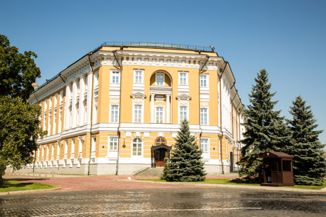 An exterior shot of a large, elegant building with a yellow and white exterior. The front and sides of the building are lined with windows and surrounded by full, tall trees.