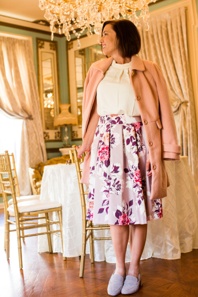 A woman in a pink coat and floral skirt standing in front of a chandelier inside Maxwell Mansion in Lake Geneva.