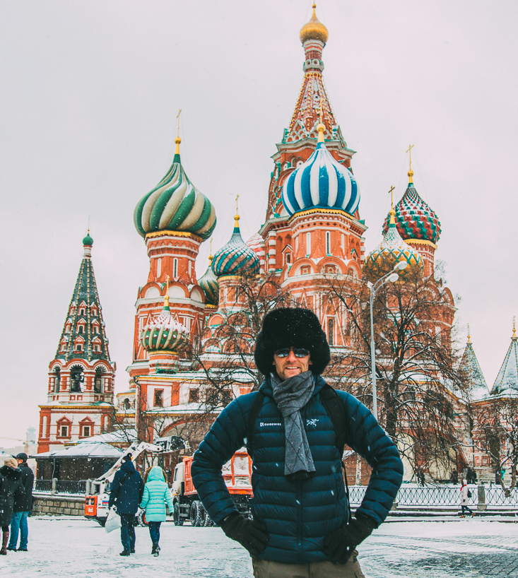 Zac poses in front of St. Basil's Cathedral in Russia on a snowy day. He's wearing a traditional Russian fur hat, puffy jacket, scarf, and gloves.