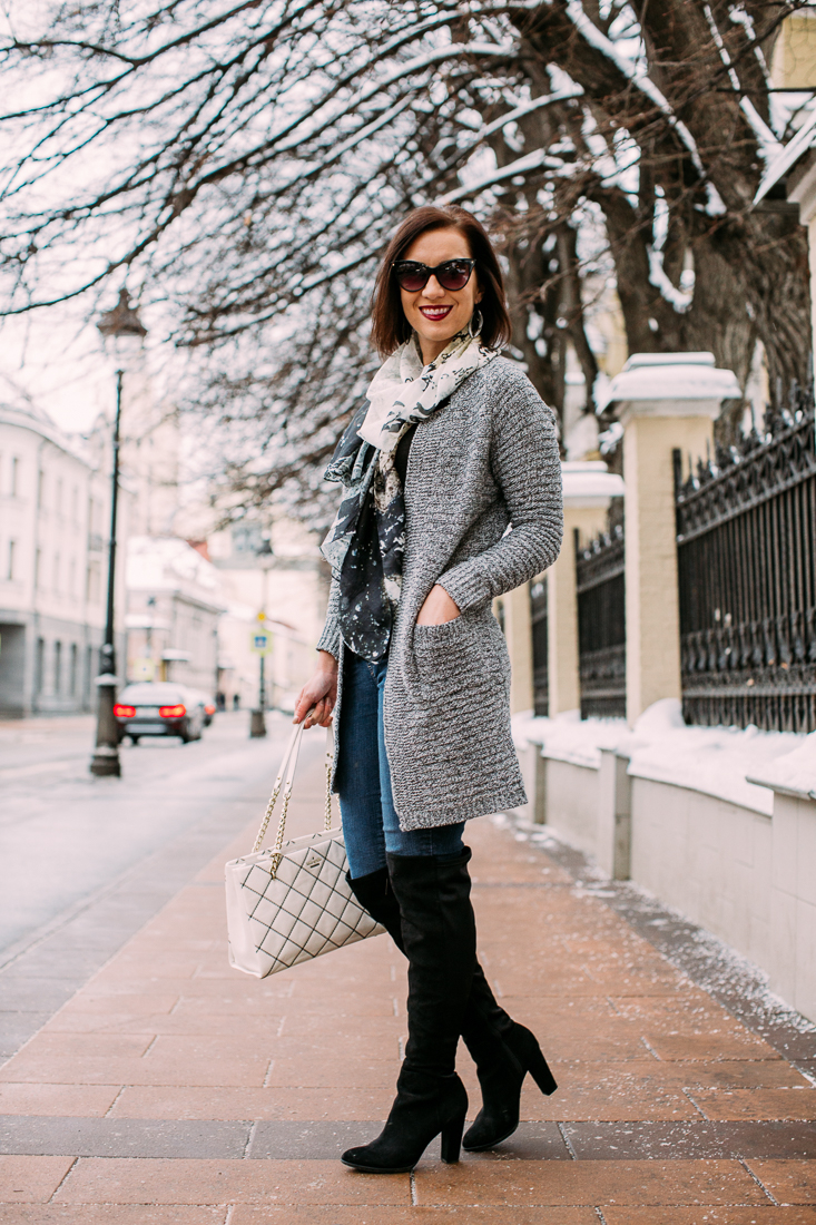 Lindsey wearing a long grey cardigan paired with black knee high boots and a black and white silk scarf and black sunglasses, she is standing on a street in Moscow during the winter