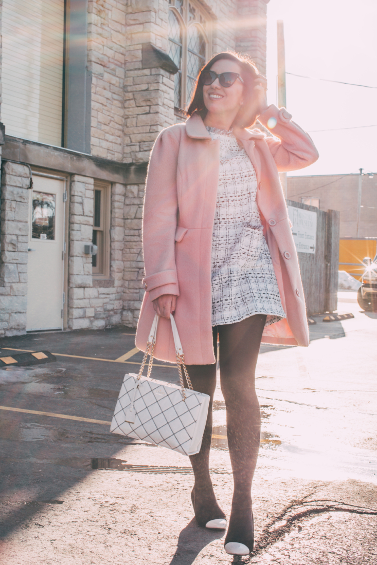 A tweed dress paired with pink coat and black tights worn by a woman with short dark hair casually touching her hair