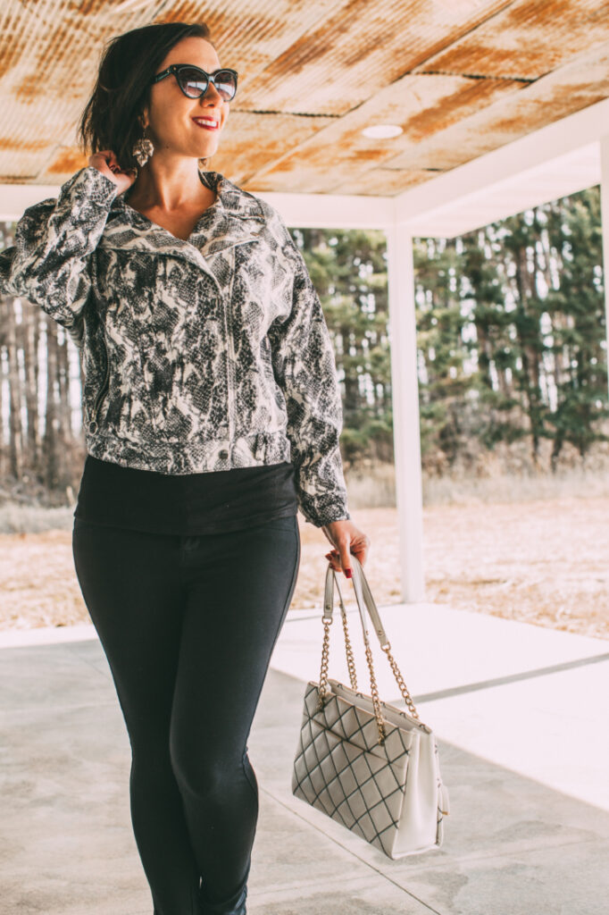 Front Pose of a women wearing snakeskin jacket, black pants, holding a white bag 