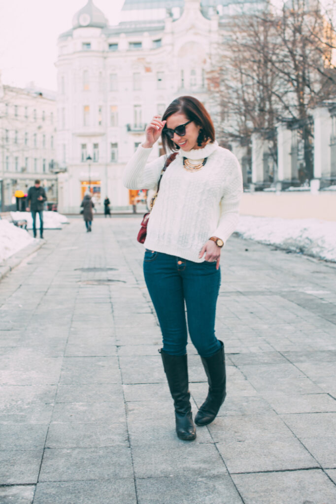 A woman poses on a sidewalk in Russia. She's wearing a white cable knit sweater, scarf, blue jeans, and calf-high boots. There's snow on the sidewalk and tree branches behind her.