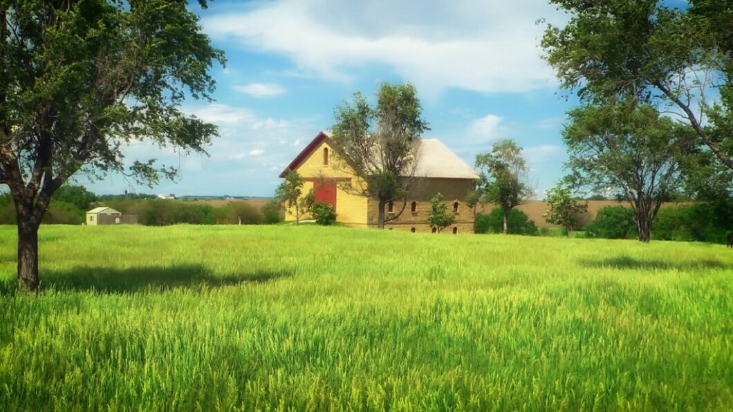 A large farmhouse sits in the distance, in the center of a large grassy field. Trees surround the landscape against a perfect blue and white cloudy sky.