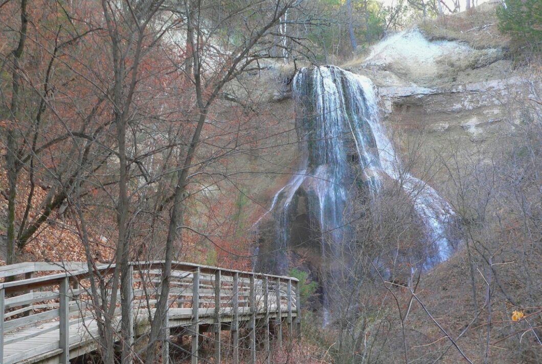 Stunning fall forestry in Nebraska! Bare trees and a ground filled with colorful fallen leaves surround a wooden footbridge, leading up to a thundering waterfall.