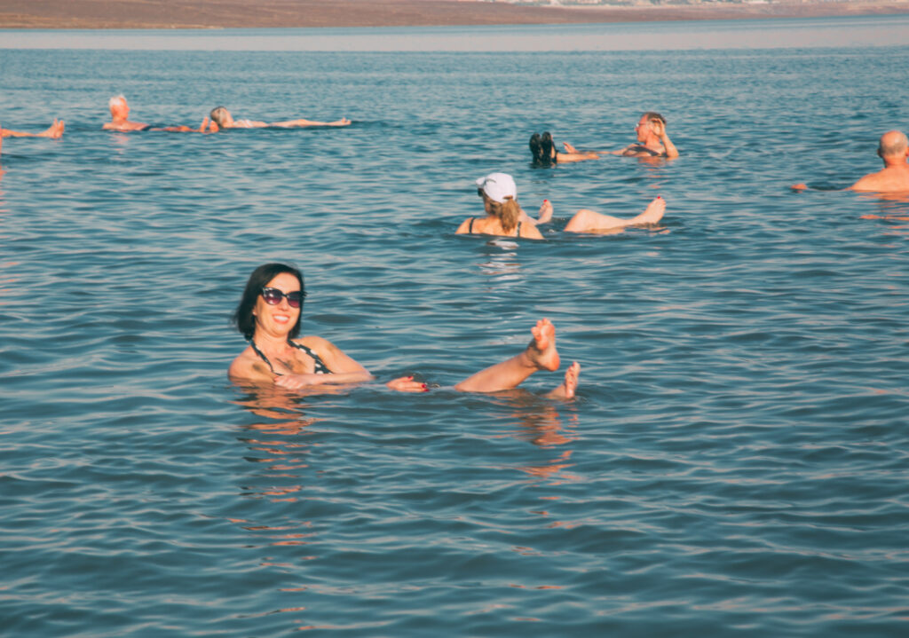 A woman with short brown hair and sunglasses floats in the Dead Sea. Her feet, shoulders, and head float above water, she appears to be sitting in the dense, salty sea water.