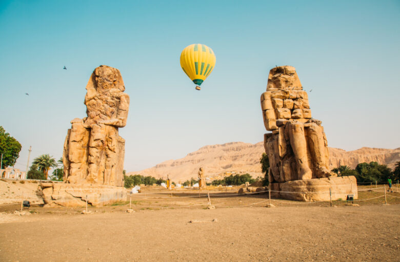 The Colossi of Memnon with a yellow hot air balloon floating between the statues.