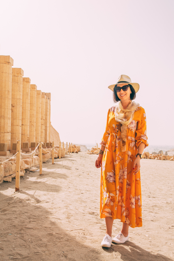 Lindsey of Have Clothes Will Travel wearing a yellow floral maxi dress, straw fedora, scarf and white keds. She's standing next to tall stone columns on a sandy walk way in Luxor, Egypt.