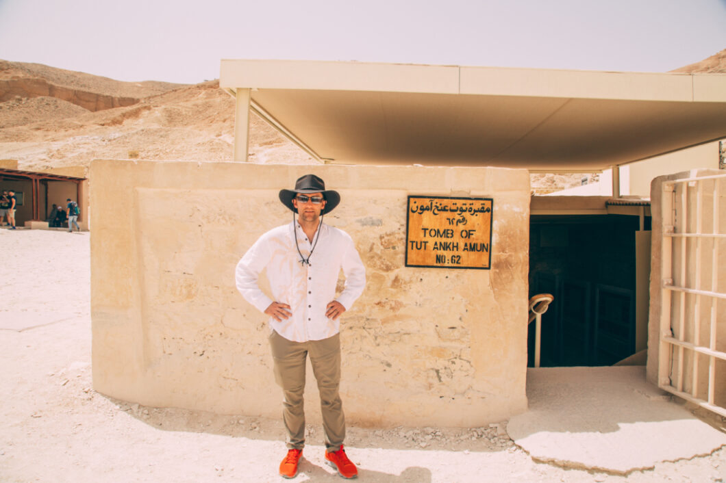 Zac wear a white linen shirt, khaki pants, bright orange sneakers and an oilskin leather hat standing in front of the entrance to King Tut's tomb with his hands on his hips.