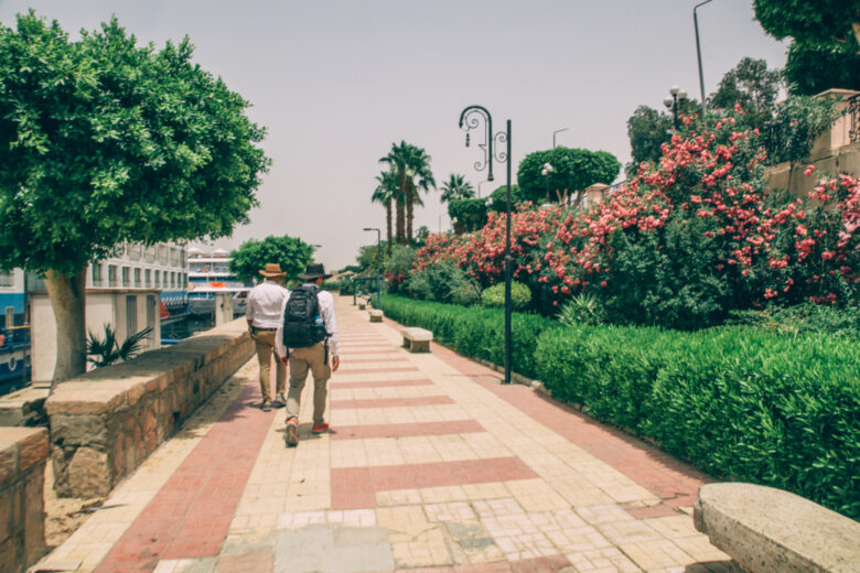 Two people wearing hats and walking ahead on a sidewalk in Luxor, Egypt/ 