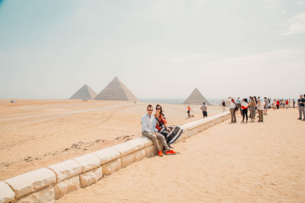 A couple sitting and posing with crowds of people photographing the Egyptian pyramids on a sunny day.