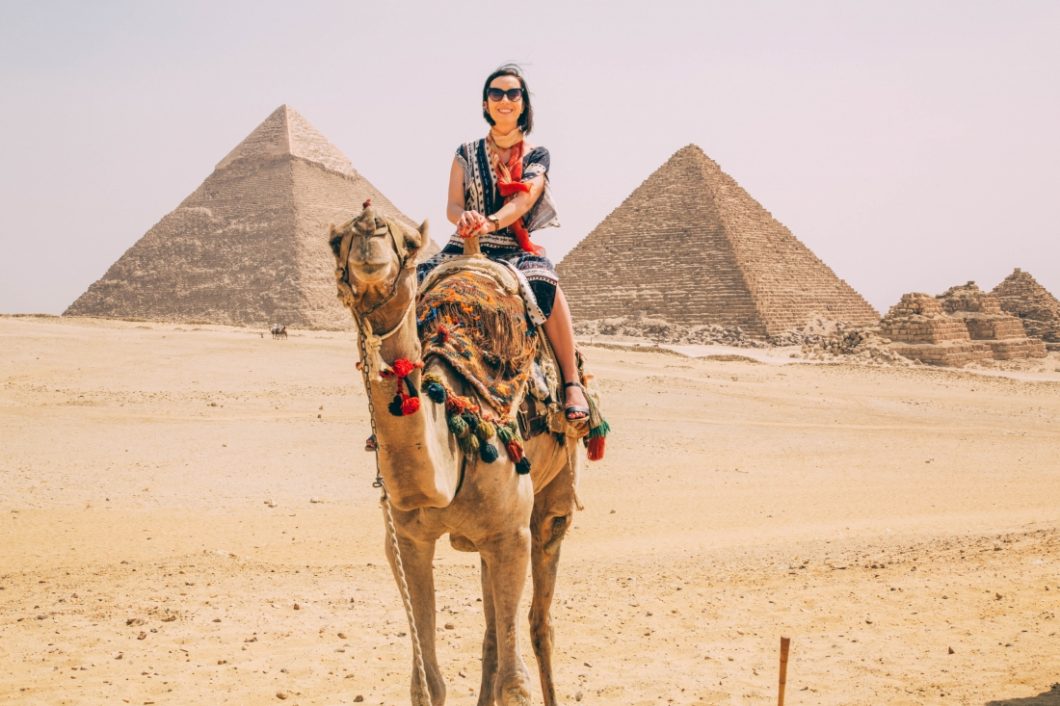 A woman riding a camel in front of the pyramids wearing a maxi dress.