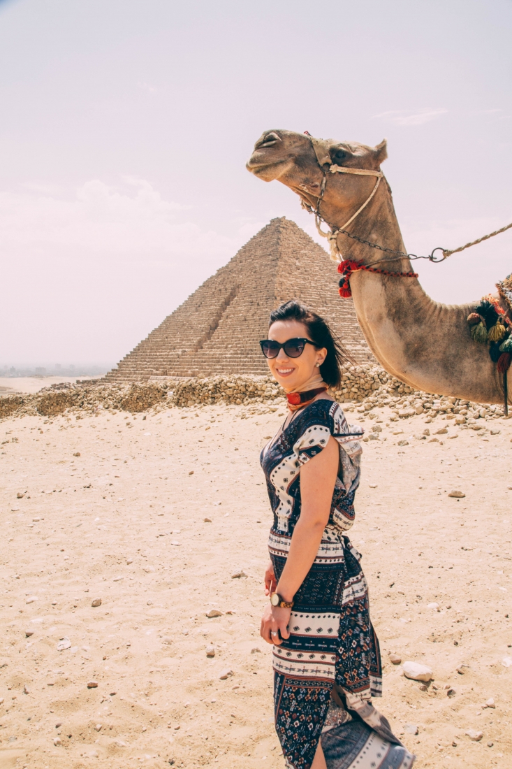 A woman in a maxi dress and sunglasses standing next to a camel in front of the pyramids of Giza.