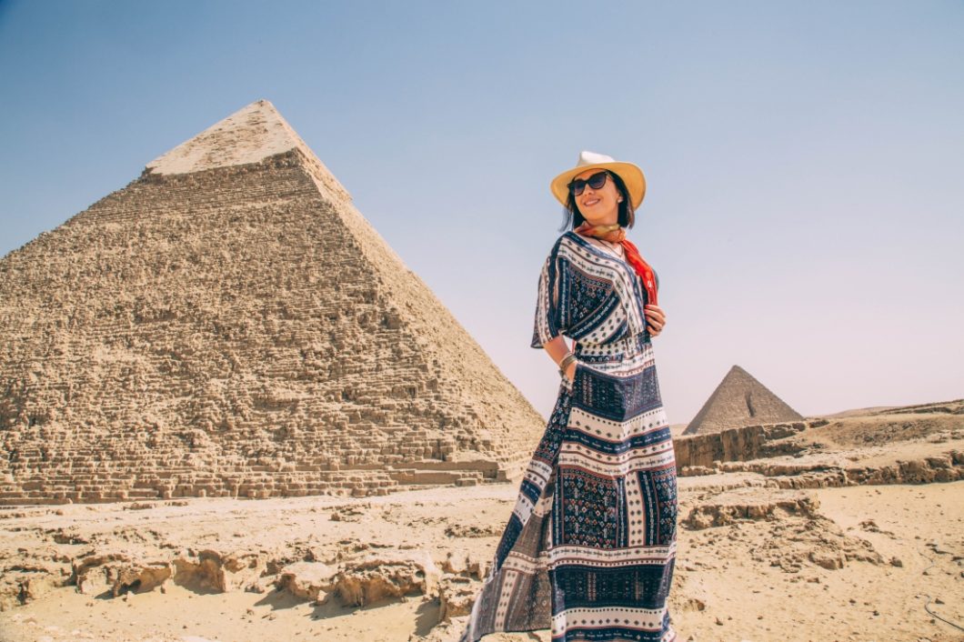 A woman is standing in front of the pyramids of Giza wearing a maxi dress, scarf, and hat.