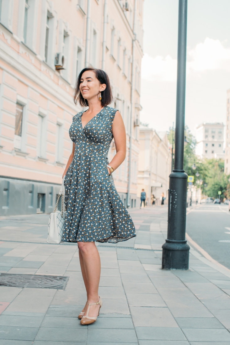 A woman poses on a city sidewalk, wearing a stylish travel dress. The dress is a light gray color with an all-over floral pattern. 