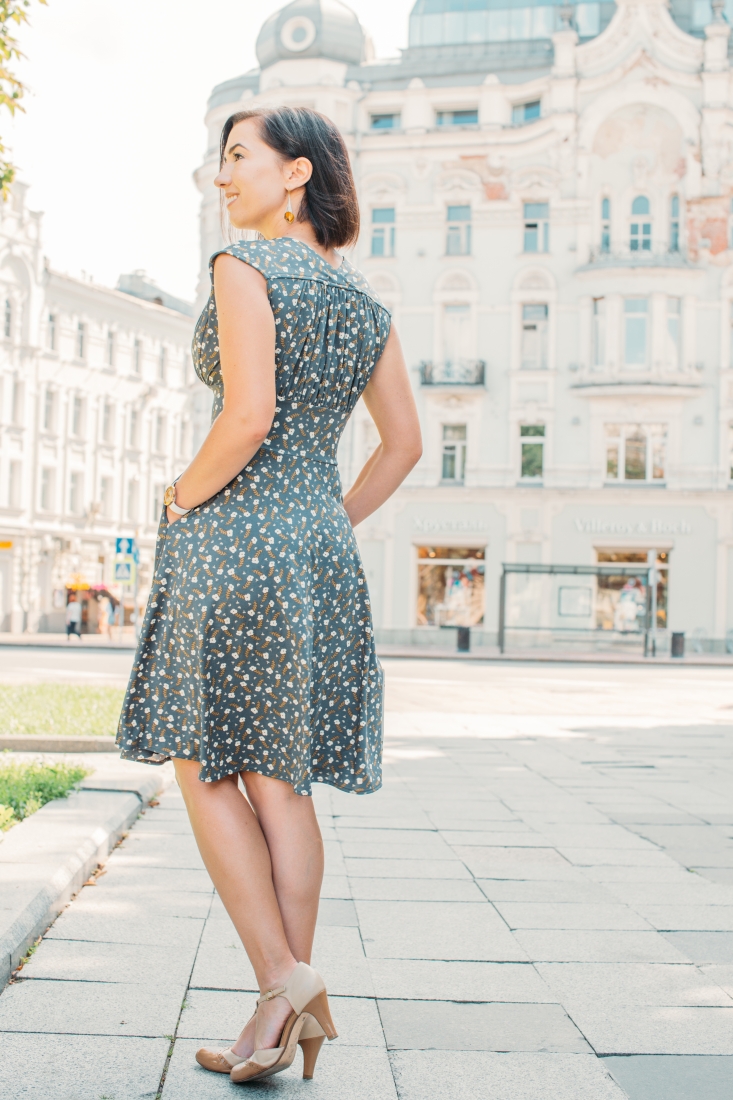 A woman poses wearing a knee-length dress with an all-over floral pattern in white, blue, and gray. Her back is towards the camera, which shows the back of the dress and her vintage style kitten heel shoes.