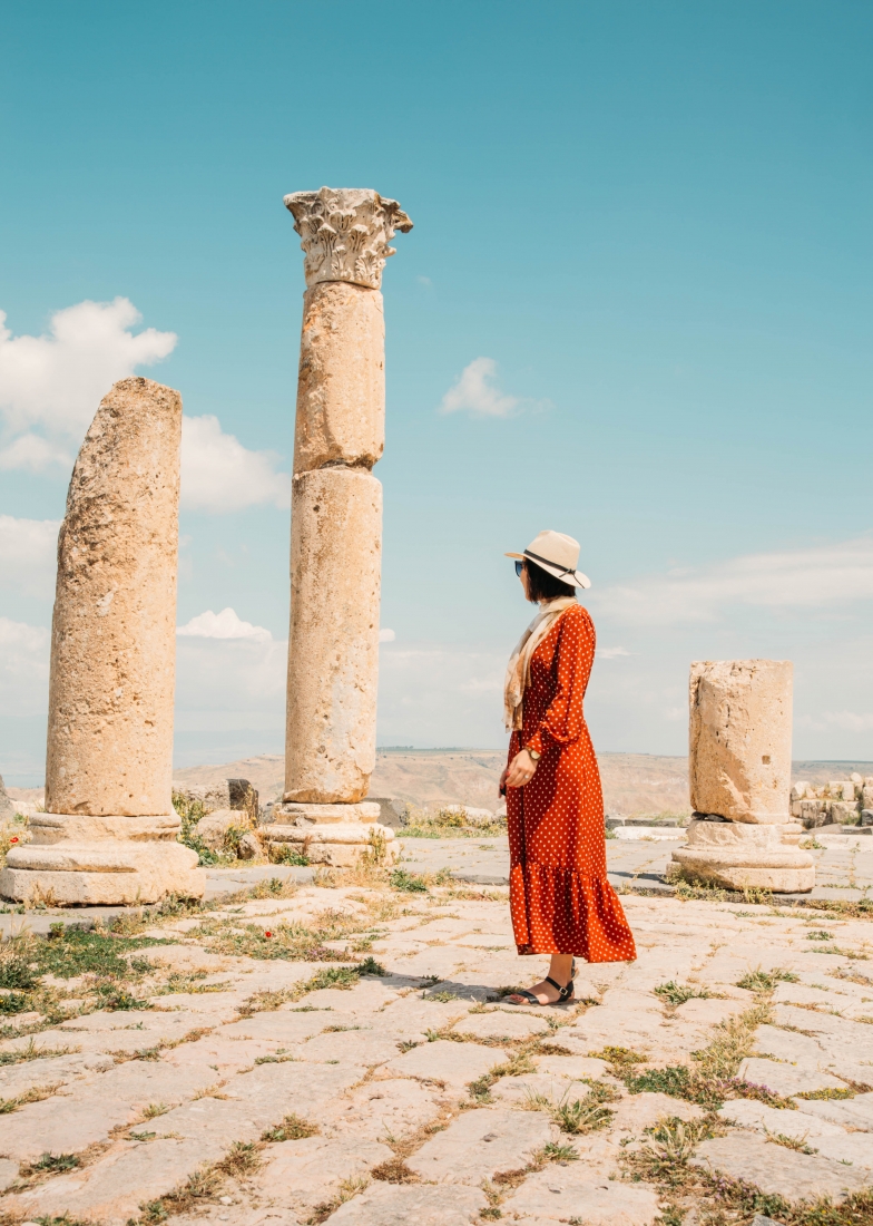 A woman poses in front of stone ruins in Jordan, facing away from the camera. She's wearing a red maxi dress with polka dots, a scarf, and sunhat and looking out over the horizon.