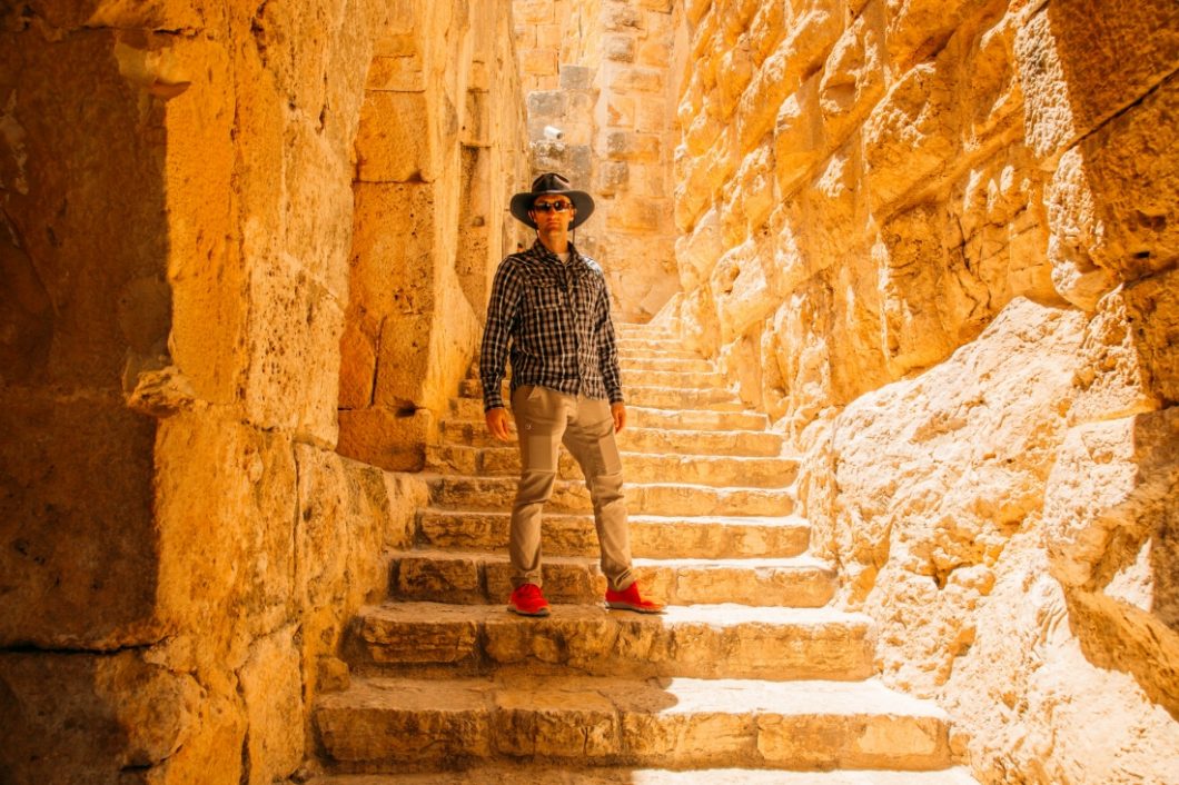 A man poses on a stone stairway between two stone walls. He's wearing khaki pants, tennis shoes, and a long sleeve plaid button-up shirt, along with a sun hat and sunglasses.