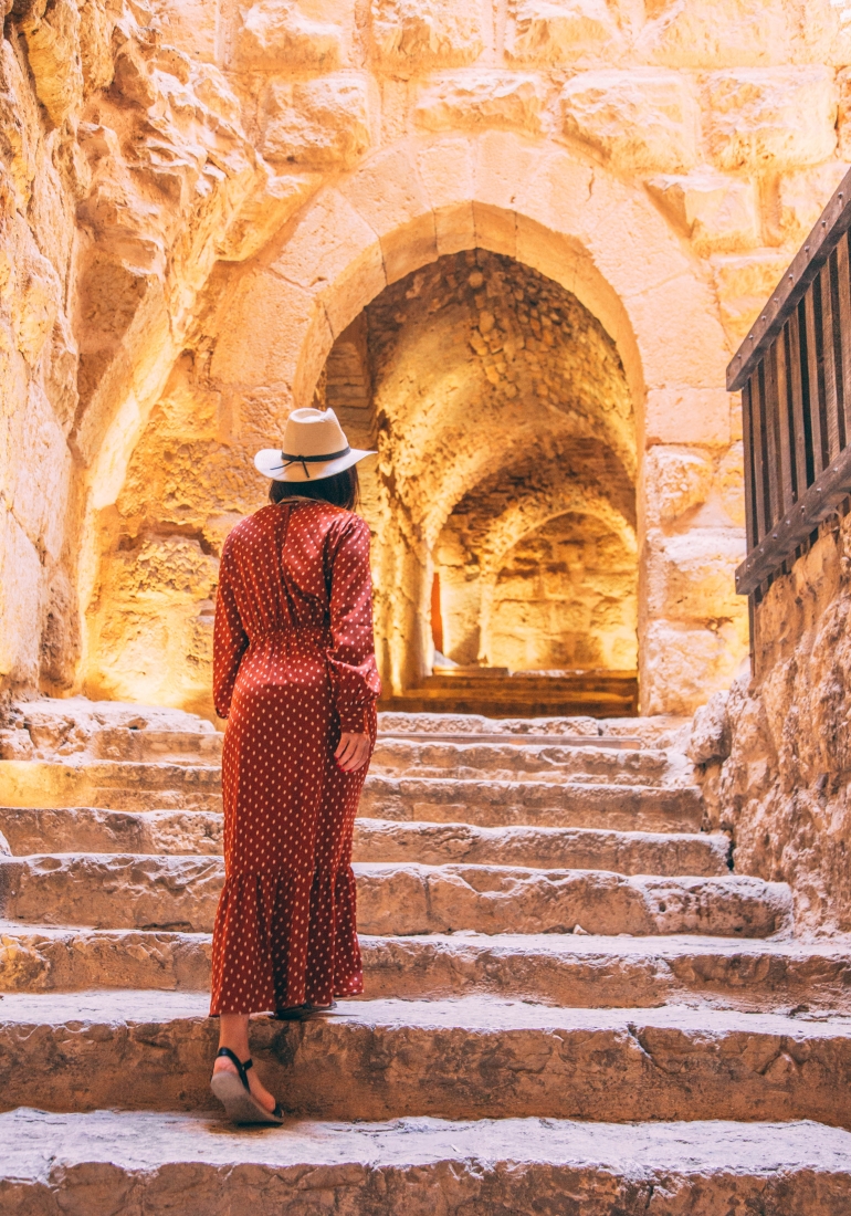 A woman walks up stone steps in Jordan, her back turned towards the camera. She's wearing a long red polka dot dress and hat.