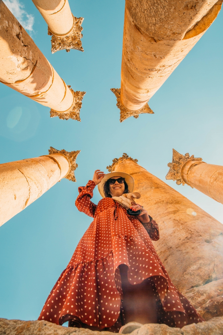 An image looking up at columns of ruins in Jerash, Jordan. A woman stands among the columns, wearing a long red dress, hat and scarf, smiling at the camera.