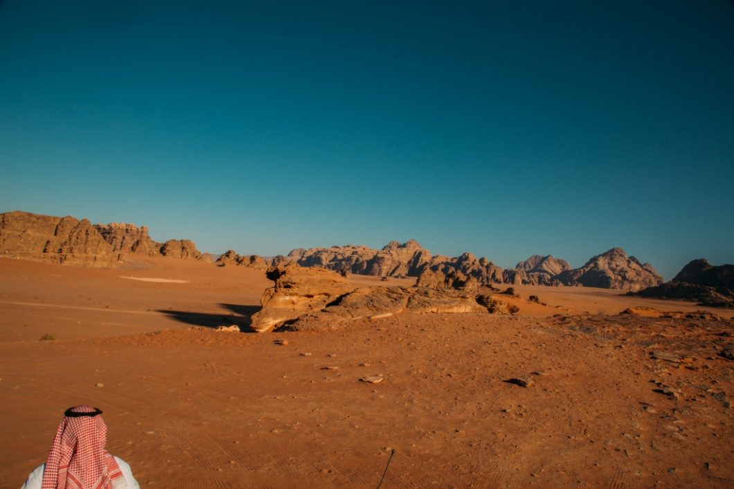 A Jordan man wearing a head scarf and white top is seen walking towards sandy, rocky cliffs in a Wadi Rum valley.