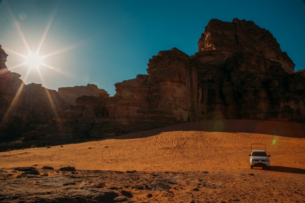 The sun shines bright over a large rocky ridge in the Wadi Rum desert. A jeep is parked in a sandy valley in front of the towering rock ridge.