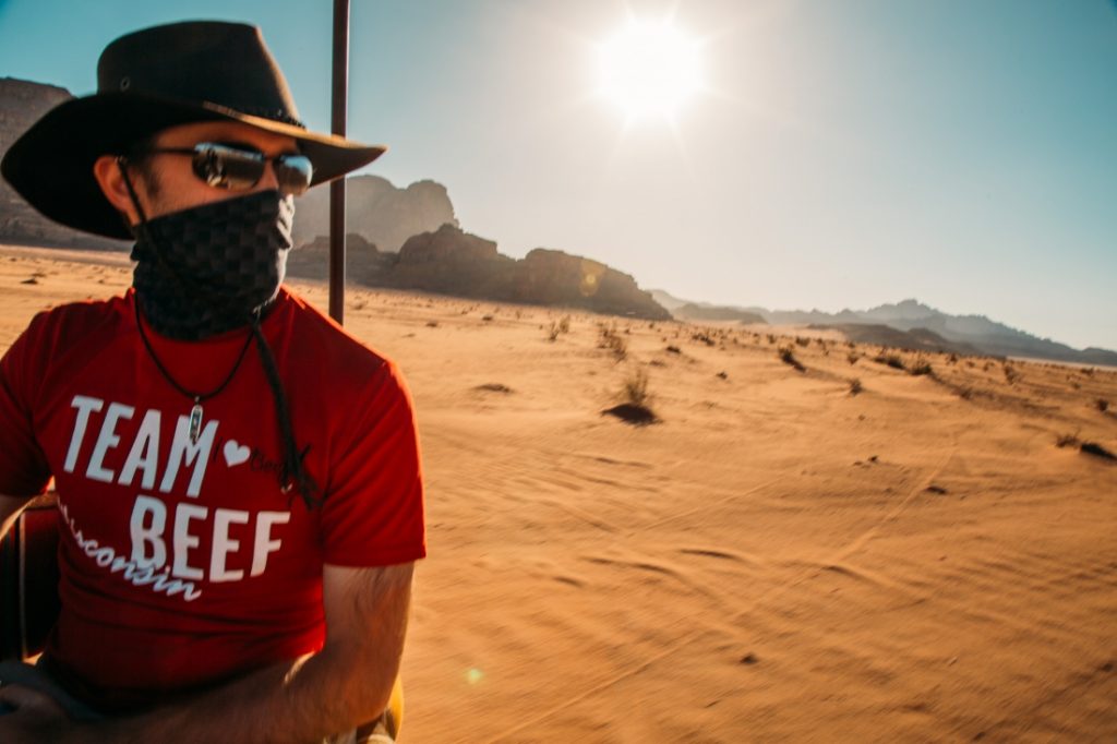 A man wearing a red shirt, hat, sunglasses, and a scarf over the lower half of his face admires the scenery in the Wadi Rum desert during a Jeep tour. Behind him, a landscape of rocky mountains and sandy valleys expands under a sunny sky.