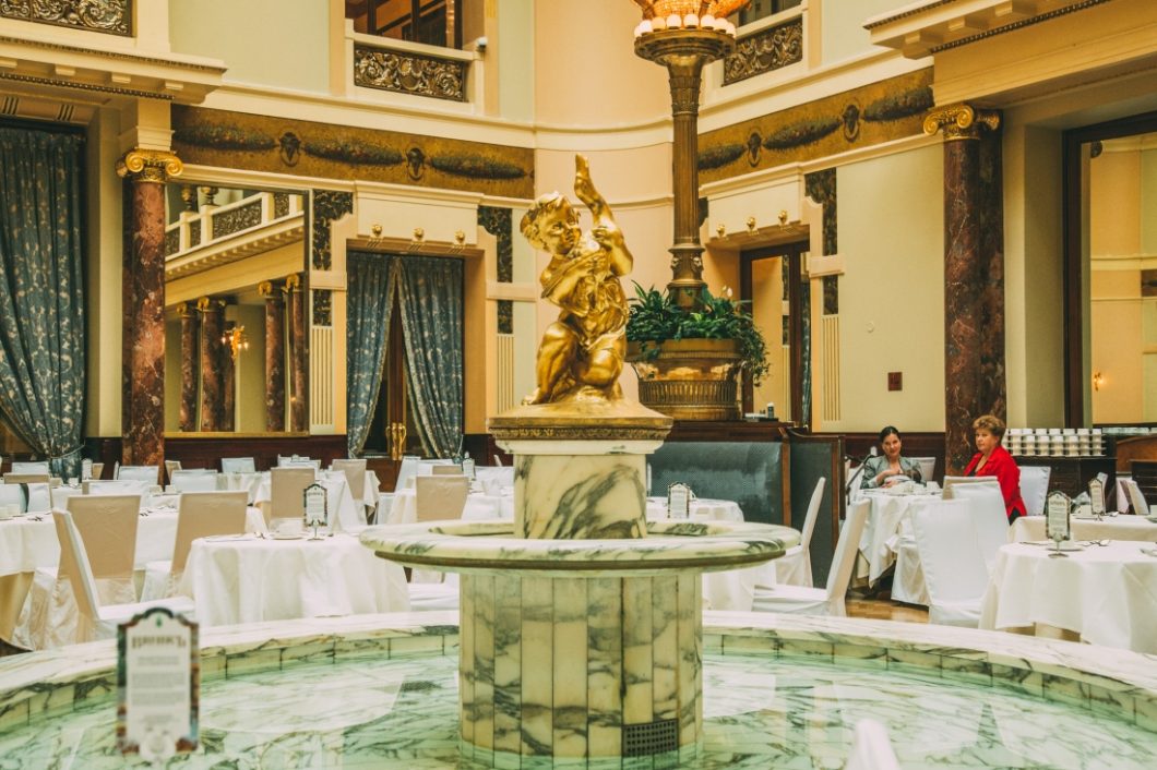 The famous fountain inside the grand dining room of the Metropol hotel in Moscow. The fountain is marble tiled with a gold statuette of an angel. White clothed tables surround the fountain in an ornate dining room.