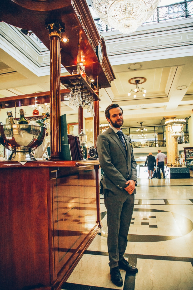 A suited Russian man named Daniel stands with his hands folded in front of a hotel bar, prepared to guide guests on a "Gentleman in Moscow" tour of the Metropol Hotel