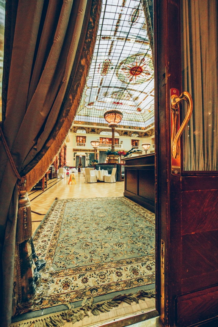 The entrance to The Piazza at the Metropol Hotel. The grand dining room lies beyond two old wood doors and features a large vaulted stained glass ceiling and many dining tables.